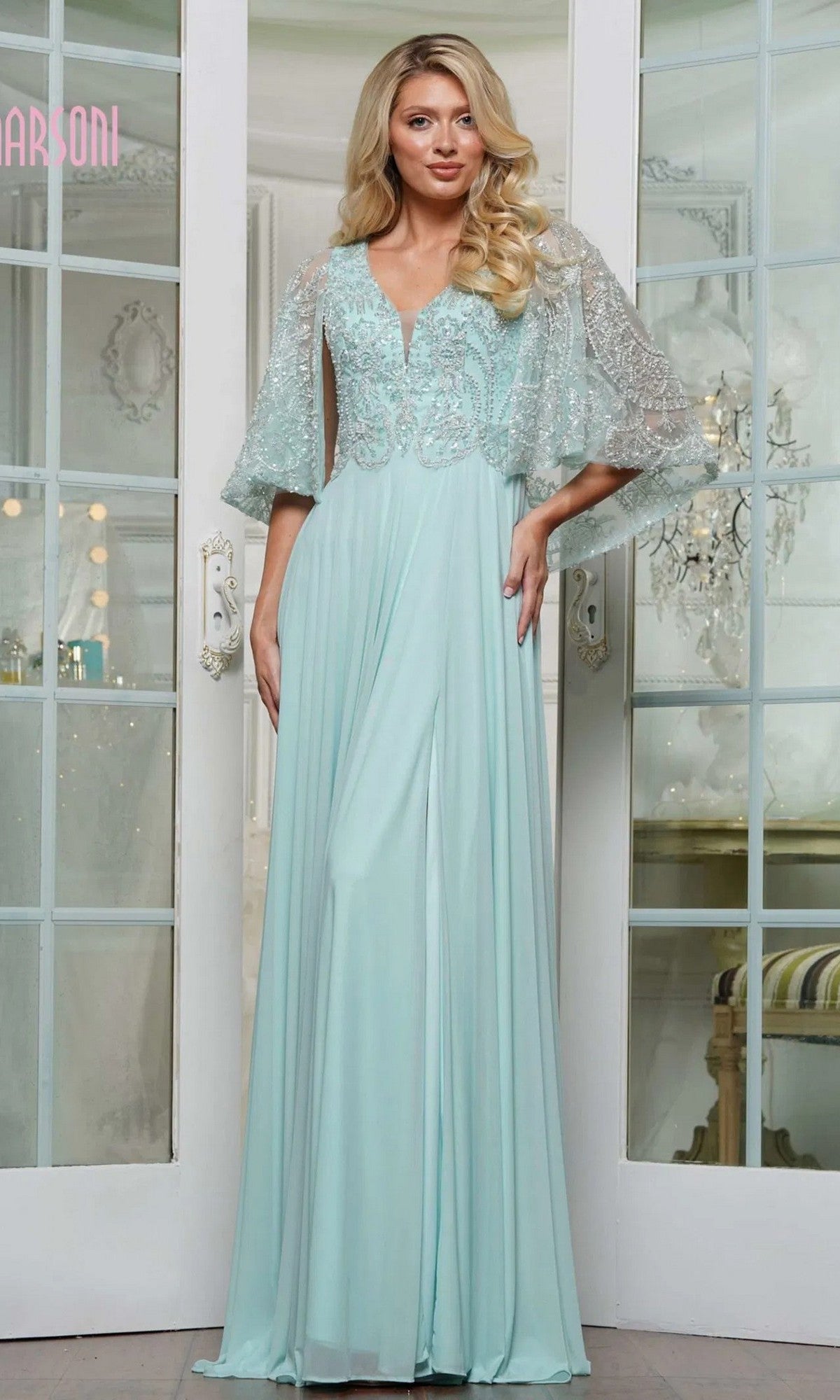 A woman poses indoors in the Marsoni MV1302, a long formal dress featuring light blue beading, lace sleeves, and intricate embroidery, in front of glass-paneled doors.